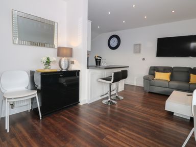 A living area with a gray sofa, yellow pillows, a wall-mounted television, two black bar stools at a counter, a black cabinet with a table lamp and flowers, and white chairs at 8 Marina View in Pwllheli