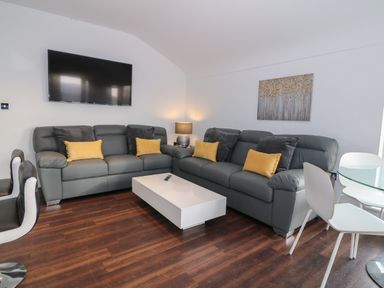A living room with gray sofas yellow and black cushions a white coffee table a wall-mounted television a lamp and a glass dining table with white chairs at 8 Marina View in Pwllheli