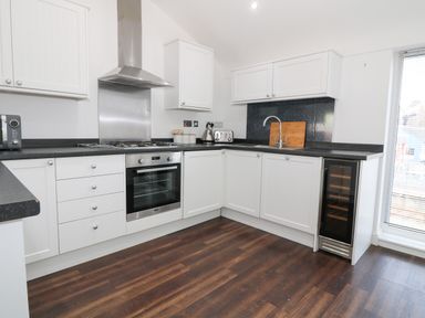 A kitchen with white cabinets black countertops a stainless steel oven and stove a sink a cutting board a kettle a toaster a wine fridge and wood flooring at 8 Marina View Pwllheli