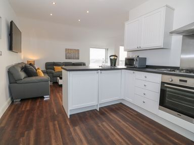 A kitchen area with white cabinets and appliances opening to a living room with grey sofas and yellow cushions at 8 Marina View Pwllheli