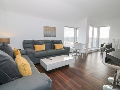 A living room with grey sofas and yellow cushions a white coffee table dark wooden floor and a dining table with white chairs at 8 Marina View in Pwllheli