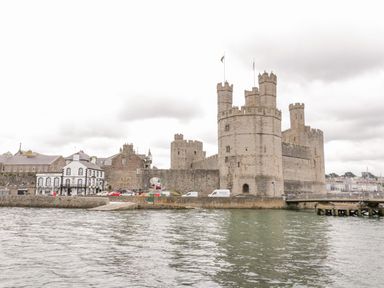 A stone castle with multiple towers next to a body of water with nearby buildings at Rhydau Cottage Pentir near Deiniolen