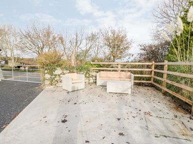 An outdoor seating area with a chair a sofa and a table on a concrete patio near a wooden fence and trees at Rhydau Cottage in Pentir near Deiniolen