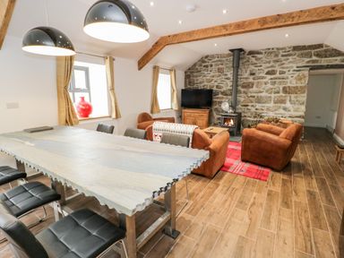A living room with a stone wall fireplace brown armchairs a dining table with black chairs and two pendant lights at Rhydau Cottage in Pentir near Deiniolen
