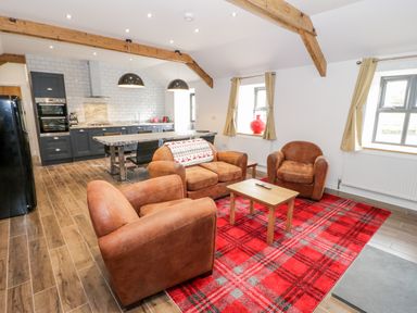 A seating area with brown armchairs and a sofa on a red plaid rug next to a kitchen with dark cabinets and two pendant lights at Rhydau Cottage in Pentir near Deiniolen