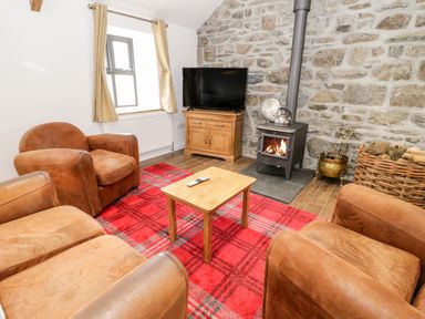 A sitting room with leather armchairs a wooden table a television on a wooden cabinet a wood stove and stone wall at Rhydau Cottage in Pentir near Deiniolen