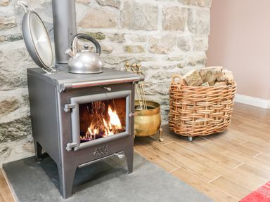 A wood burning stove with a kettle on top next to a wicker basket filled with firewood and a brass container against a stone wall at Rhydau Cottage in Pentir near Deiniolen