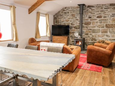 A living room with brown armchairs a stone wall with a wood stove a television on a wooden stand and a wooden dining table at Rhydau Cottage Pentir near Deiniolen