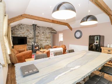 A living room with a stone fireplace a wood stove and brown chairs a television a clock on the wall and a dining table with chairs at Rhydau Cottage in Pentir near Deiniolen
