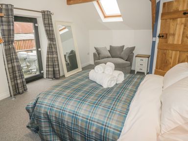 A bedroom with a bed covered with a checkered blanket and white towels on top a gray armchair a white nightstand a full-length mirror a wooden door and a glass door with plaid curtains at Rhydau Cottage in Pentir near Deiniolen
