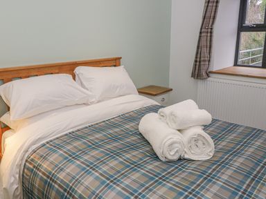 A bedroom with a wooden bed frame white pillows rolled white towels on a plaid bedspread near a window at Rhydau Cottage in Pentir near Deiniolen
