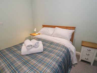 A bedroom with a bed with plaid bedding white pillows a small lamp on a bedside table and rolled towels on the bed at Rhydau Cottage in Pentir near Deiniolen