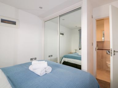 A bedroom with a bed covered by a blue blanket folded white towels on the bed a mirrored wardrobe and an open door leading to a bathroom at 28 Zenith in Porth