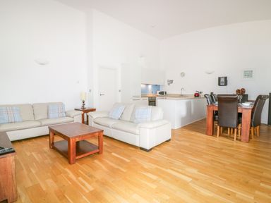 A living room with two cream sofas and a wooden coffee table next to a kitchen with white cabinets and a dining table with six dark chairs at 28 Zenith in Porth