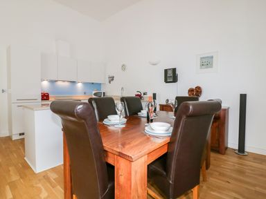 A dining area with a wooden table and six chairs set with plates glasses and a bottle with a kitchen in the background at 28 Zenith in Porth
