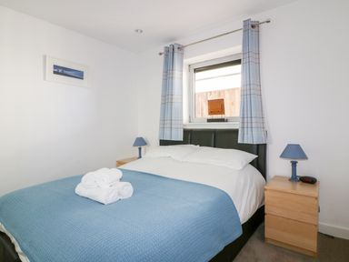 A bedroom with a bed with blue blanket and white sheets two bedside tables with blue lamps and a window with blue curtains at 28 Zenith in Porth