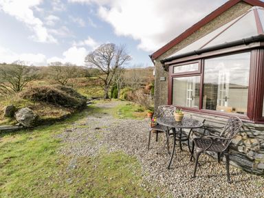 An outdoor seating area with a metal table and two chairs next to a building and a gravel pathway with trees and grass at Melin Newydd in Porthmadog