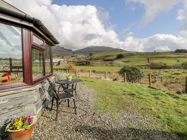 An outdoor seating area with a small table and two chairs next to a stone wall window overlooking grassy fields and hills at Melin Newydd in Porthmadog