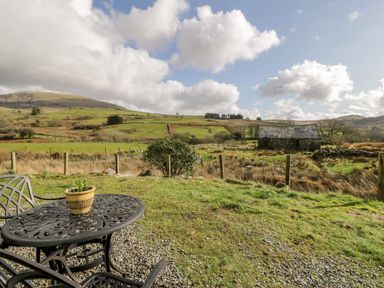 An outdoor area with a metal table and chairs with a potted plant and a view of green fields stone building and hills at Melin Newydd in Porthmadog
