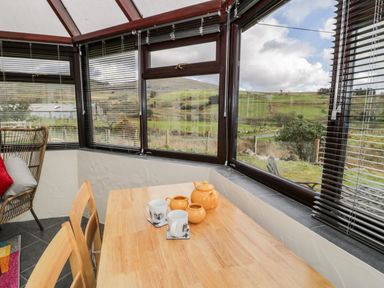 A dining area with a wooden table and chairs next to windows showing a rural landscape at Melin Newydd in Porthmadog