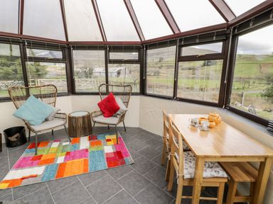 A sunroom with two wicker chairs with cushions colorful rug a small table and a wooden dining table with chairs at Melin Newydd in Porthmadog