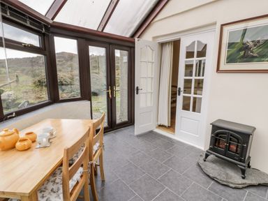A dining area with a wooden table and chairs tea set on the table glass doors and windows and a small electric stove at Melin Newydd in Porthmadog