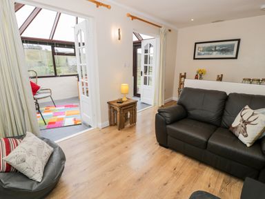 A living room with black leather sofas and a small wooden table with a lamp next to an open door leading to a glass sunroom at Melin Newydd in Porthmadog