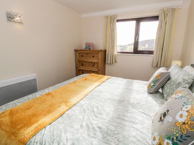 A bedroom with a bed with floral bedding and embroidered cushions a wooden chest of drawers and a window with curtains at Melin Newydd in Porthmadog