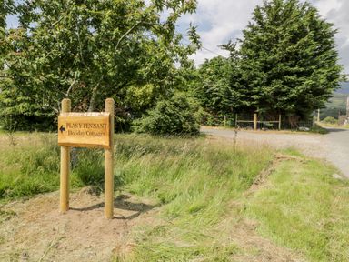 A wooden sign pointing to Plas Y Pennant holiday cottages near a grassy path with trees and a country road