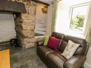 A living room with a brown leather sofa with three cushions next to a stone wall and a window with green curtains at Bwthyn Pennant in Porthmadog