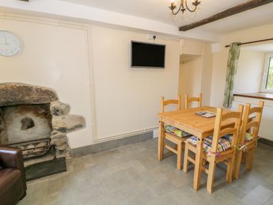 A dining area with a wooden table and four chairs next to a stone fireplace and a wall-mounted television at Bwthyn Pennant in Porthmadog
