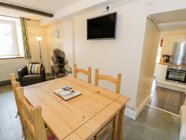 A dining room with a wooden table and chairs a television on the wall an armchair a fireplace and a view into a kitchen at Bwthyn Pennant in Porthmadog