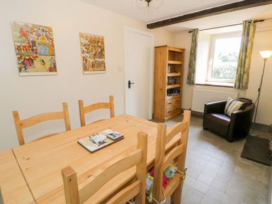 A dining room with a wooden table and chairs a bookcase an armchair with a pillow and a window with green curtains at Bwthyn Pennant in Porthmadog