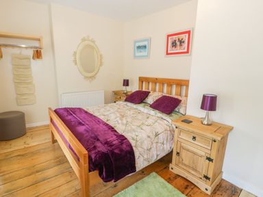 A bedroom with a wooden bed with purple pillows and blanket two wooden bedside tables with lamps a mirror and two framed pictures on the wall at Bwthyn Pennant in Porthmadog