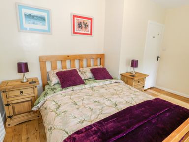 A bedroom with a wooden bed frame, floral bedding, two purple pillows, two wooden bedside tables with purple lamps, and two framed pictures on the wall at Bwthyn Pennant in Porthmadog