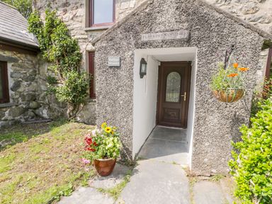 Stone house entrance with a wooden door a flower pot on the ground and a hanging flower basket at Bwthyn Pennant in Porthmadog