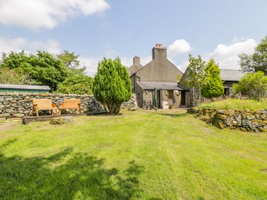 A grassy backyard with wooden benches stone walls and a stone house at Bwthyn Pennant in Porthmadog