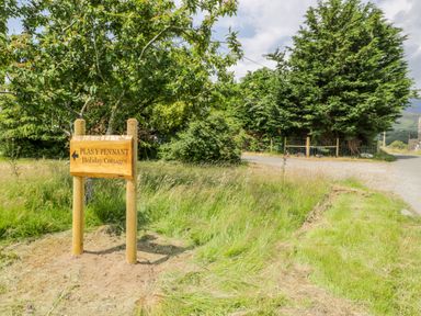 A wooden signpost for Plas y Pennant Holiday Cottages near a grassy roadside with trees in the background at Bwthyn Pennant in Porthmadog
