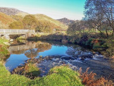 A river with a small waterfall and stone bridge with white railings in a rural landscape at Bwthyn Pennant in Porthmadog