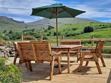 An outdoor wooden table set with benches and chairs under a green umbrella on a deck with green hills in the background at Bwthyn Pennant in Porthmadog