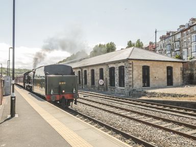 A steam train at a railway station with a stone building and residential houses in the background at Premier Transit & Lady Hilda at Engine Shed in Whitby