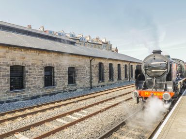 A black steam locomotive on railway tracks next to a stone building with arched windows at Premier, Transit & Lady Hilda @ Engine Shed in Whitby