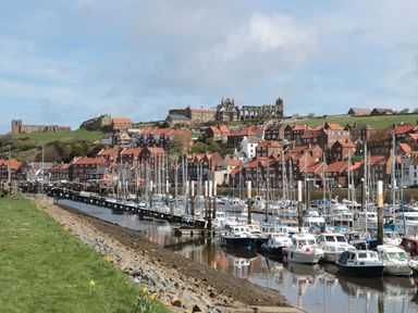 A marina with boats docked alongside a shoreline with grass and rocks and a town with red-roofed buildings and ruins on a hill in the background at Premier Transit & Lady Hilda @ Engine Shed in Whitby