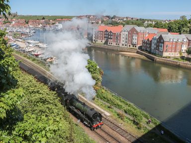 A steam train traveling on railway tracks beside a river with houses and boats in the background at Premier Transit and Lady Hilda Engine Shed in Whitby
