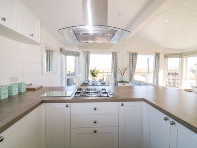 A kitchen with stove and extractor fan looking towards a dining area with table set and windows with curtains at Ash Lodge in Teigngrace near Newton Abbot