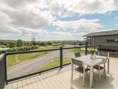An outdoor deck with a table and four chairs overlooking a gravel road and green landscape at Ash Lodge in Teigngrace near Newton Abbot
