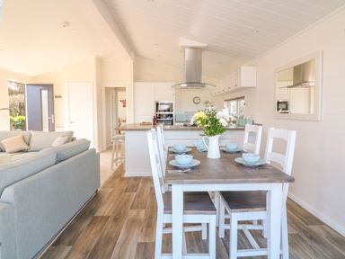 A dining area with a wooden table set with bowls and a flower vase next to an open plan kitchen and a seating area at Beech Lodge in Teigngrace near Newton Abbot
