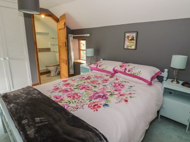 A bedroom with a bed with floral bedding two bedside tables with lamps a window and an open door to a bathroom at Cottage on the Common in Westbury-on-Severn