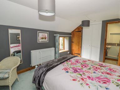 A bedroom with a bed with floral bedding a wicker chair a wall mirror and a door leading to a bathroom at Cottage on the Common in Westbury-on-Severn