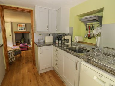 A kitchen with white cabinets, a sink, a dishwasher, utensils hanging on a rack, and a view into a living room with a fireplace at Cottage on the Common in Westbury-on-Severn